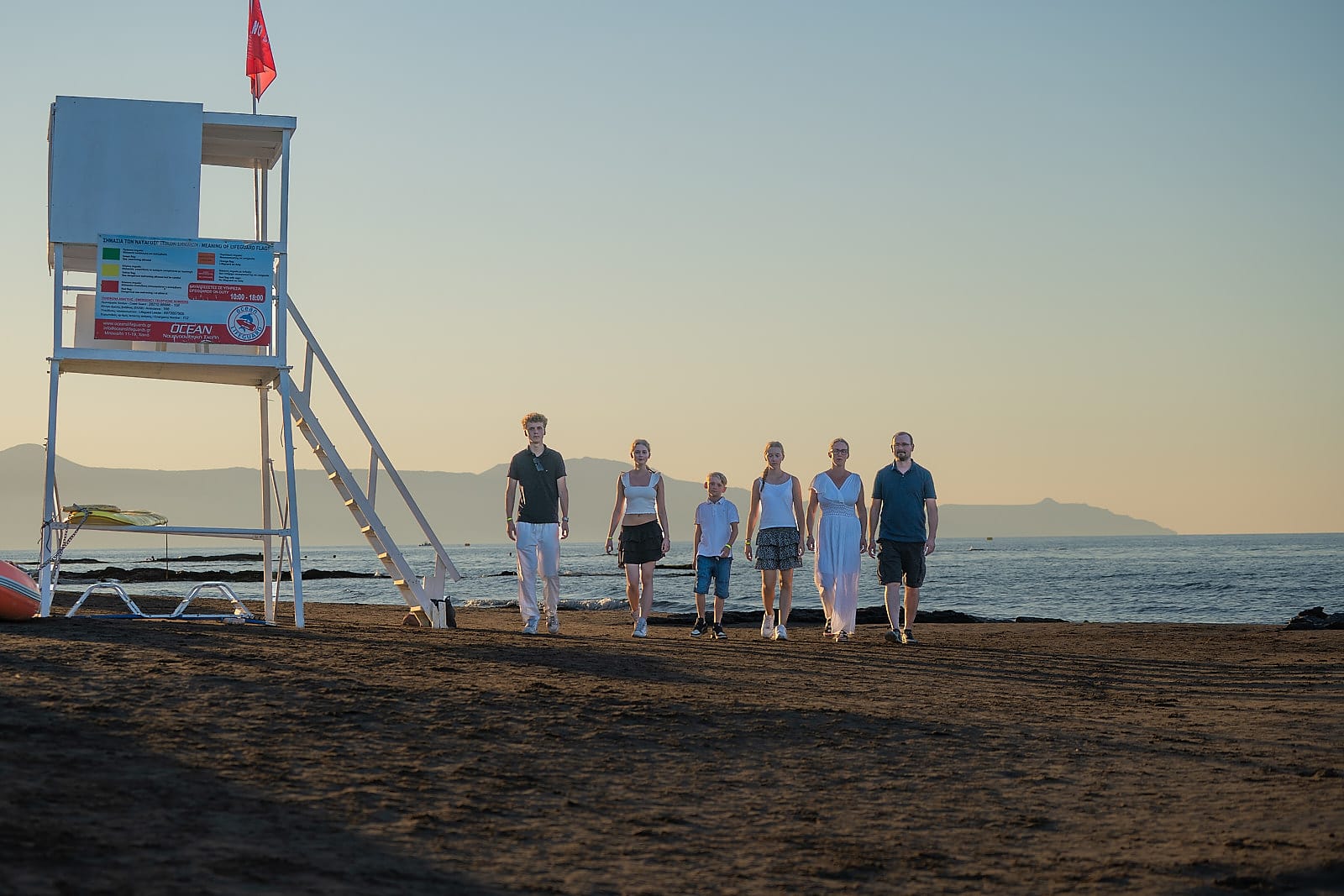 Family walking along Agia Marina beach past a lifeguard tower at sunset