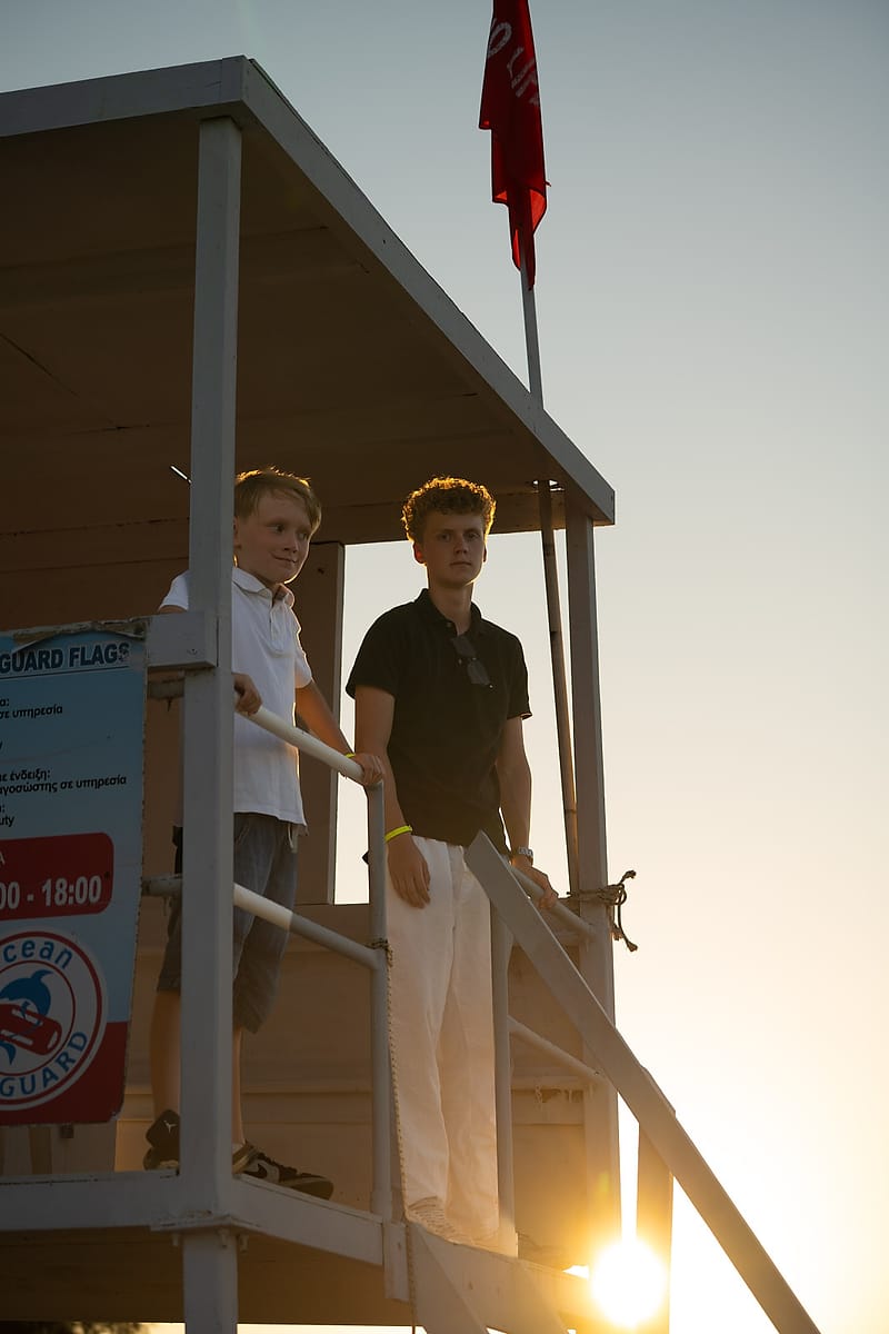 Two brothers standing on a beach lifeguard tower, lit by the setting sun
