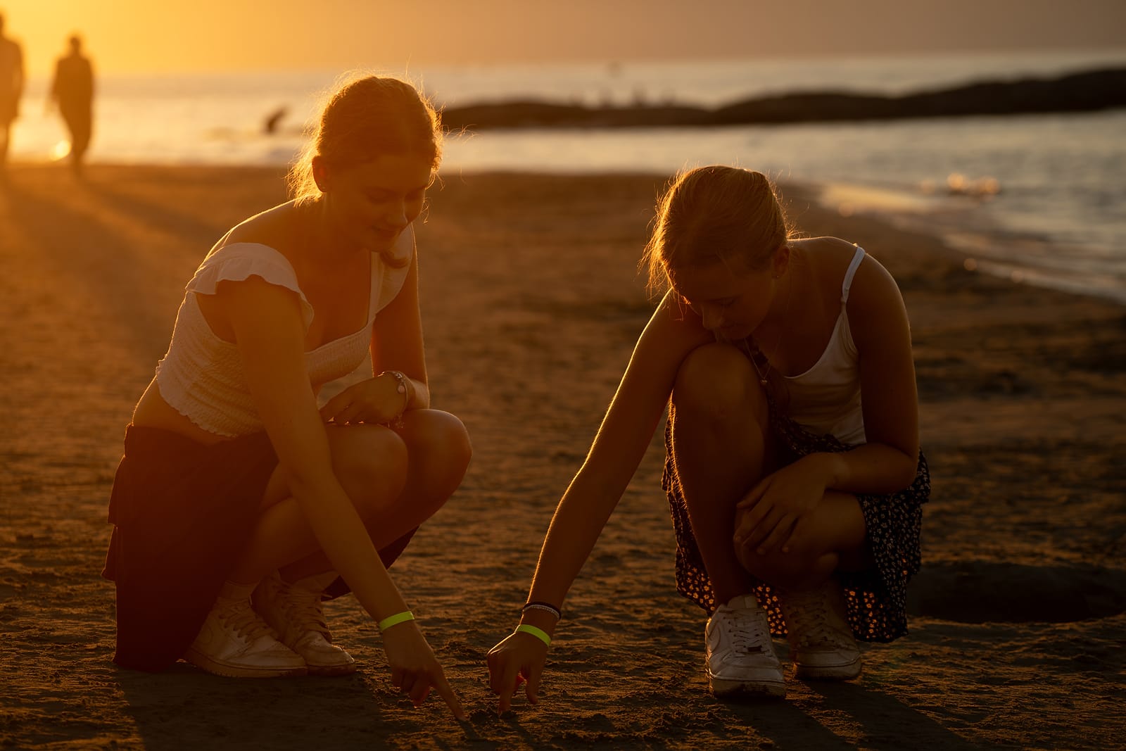 Two young women crouching to draw in the sand, backlit by the low evening sun